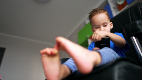 little caucasian toddler sits looking focused on the phone. cute kid on the chair his bare feet to camera. low angle view.