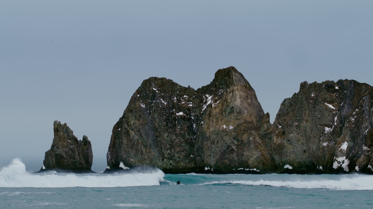 Klippefyldte havstabler stiger op fra havet med brændende bølger, naturlig baggrund med lille surfer langs skummet, Kamchatka Rusland, slow motion.