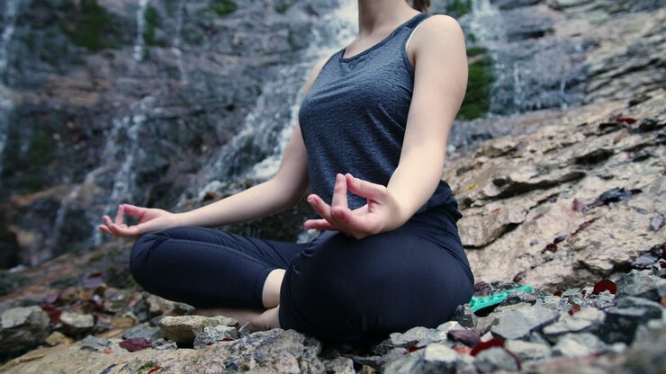 Meditación en la naturaleza, una chica relajándose en una postura de yoga cerca de una cascada, cámara en mano.