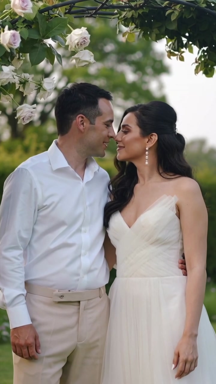 Vertical video: Photog cueing couple leaning in for near-kiss in garden, floral arch, wedding dress
