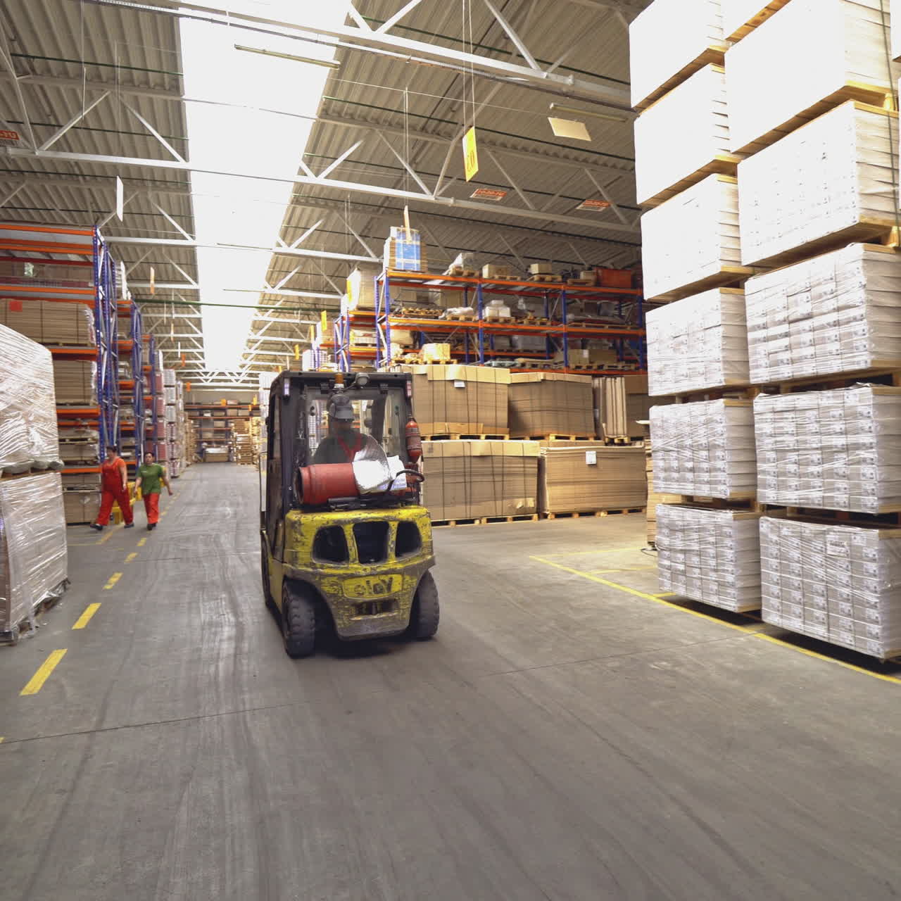 Operator in a helmet drives a yellow electric forklift through the warehouse in search of the right product on the background of huge shelves.