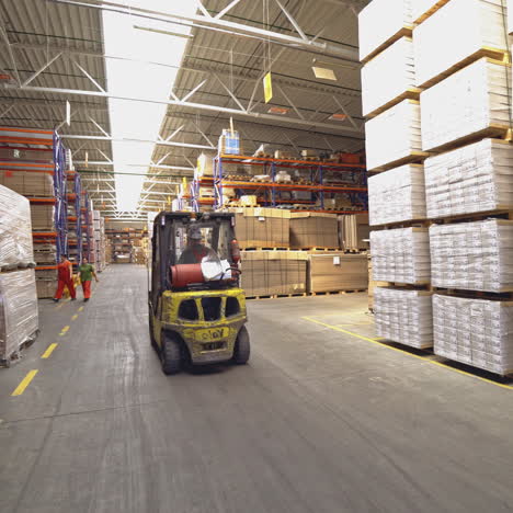 operator in a helmet drives a yellow electric forklift through the warehouse in search of the right product on the background of huge shelves.