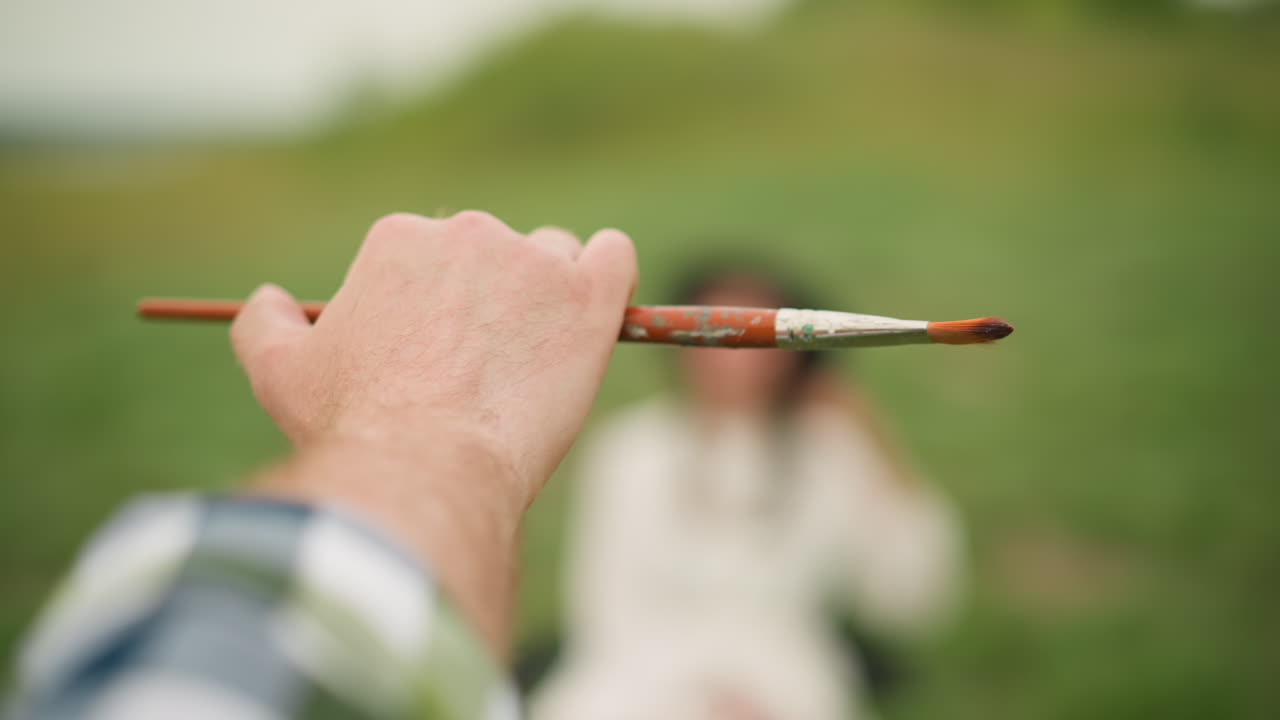 Detailed close-up of a painter's hand holding a small brush with a blurred person in the soft green background, highlighting artistic focus