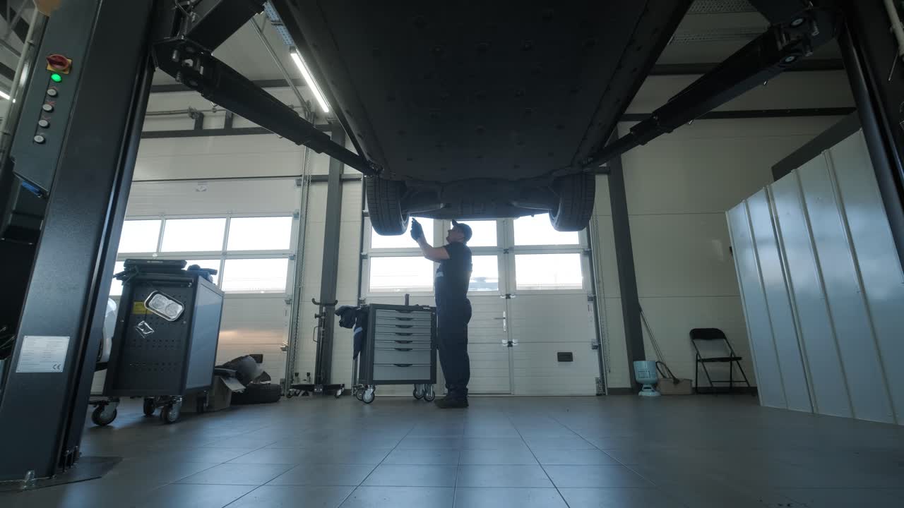 Meccanico in uniforme blu ispeziona e ripara la parte inferiore di un veicolo sollevato in un'officina professionale. L'officina è ben illuminata e attrezzata con strumenti