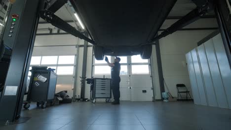 auto mechanic in a blue uniform inspecting and repairing the underside of a lifted vehicle in a professional repair shop. the workshop is well-lit and equipped with tools