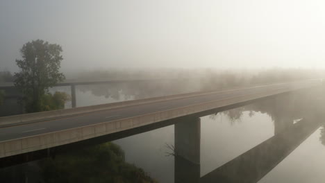 heavy fog hangs over bridge crossing calm river with early morning sunlight shining through