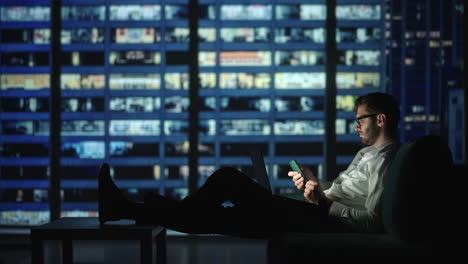 look into the screen of a mobile phone against the background of a large night city in a large window. overlooking the big city a man lies on a soph and looks at the screen of a young phone.