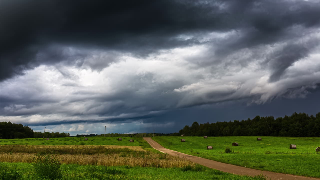 Shot of mystic dark clouds flying over green field with hay bales beside a gravel pthway in timelapse