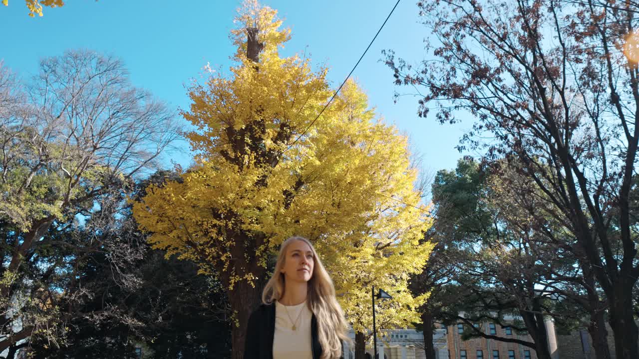 A woman strolls peacefully through a serene garden in Tokyo, surrounded by vibrant yellow autumn foliage.