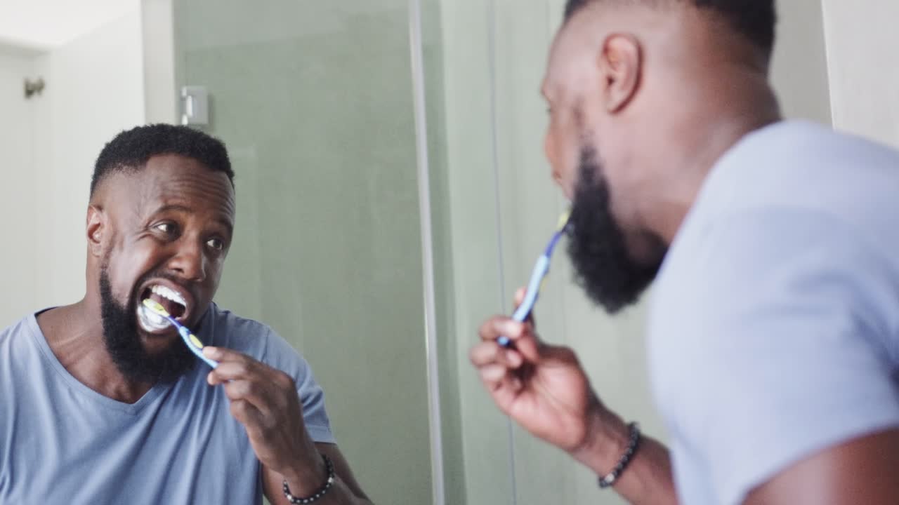 African American man brushing teeth at bathroom sink, watching reflection with blue toothbrush