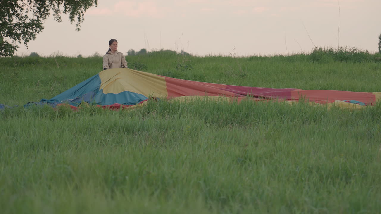 close up of tall grass framing woman kneeling in field pulling vibrant hot air balloon envelope open with gloved hands during preflight balloon preparation in rural sunset landscape
