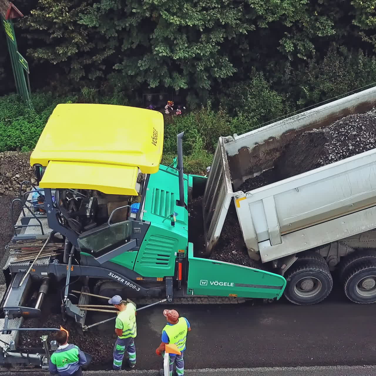A process of laying asphalt by repairmen using an asphalt paver, a vibro-roller and a truck on a highway between cities. Aerial view.