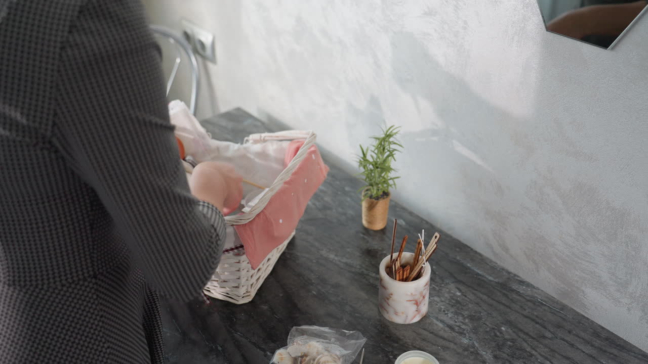 Side view of white girl in checkered dress unpacking shopping basket on marble table, carefully removing groceries and placing them beside small potted plant and jar of sticks