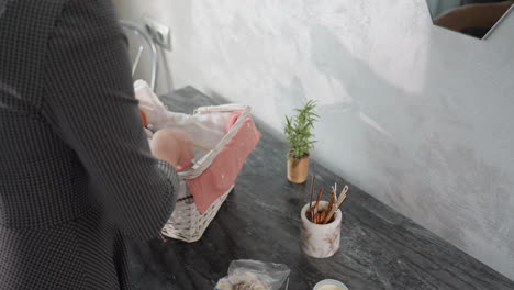 side view of white girl in checkered dress unpacking shopping basket on marble table, carefully removing groceries and placing them beside small potted plant and jar of sticks