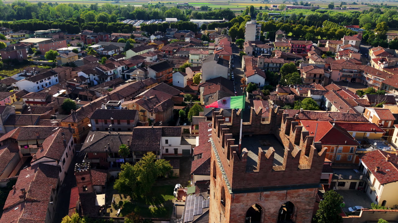 Drone orbit around a medieval stone tower with a local flag, revealing historic rooftops and dense urban fabric in a Piedmont town.