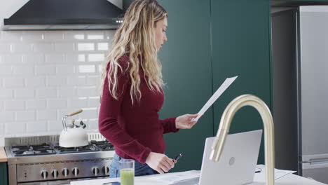 biracial woman using laptop and holding documents in kitchen at home, slow motion