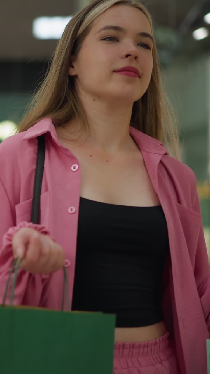 dama de vestido rosa y pantalones cortos sosteniendo bolsas de compras rojas, verdes y verdes con una cálida sonrisa, caminando a través de un centro comercial brillantemente iluminado con reflejos de luces en el vidrio en el fondo