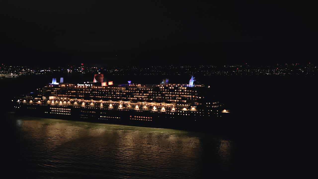 The Queen Mary 2 ocean liner sailing out of Southampton harbour at night with the city lights in the background pull out drone shot