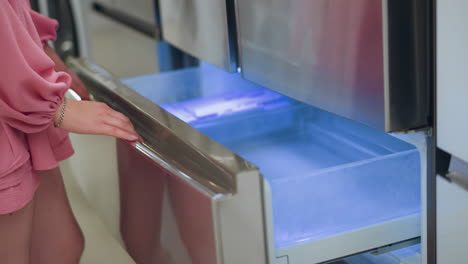 lady wearing pink dress opens lower refrigerator drawer illuminated by soft blue light in a brightly lit store, her hand, wearing a silver bangle, holds the handle while exploring the appliance