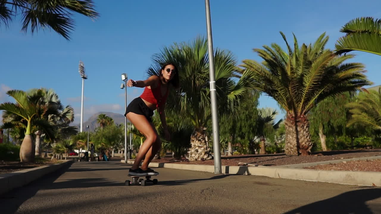 Two young Hispanic women leisurely skateboard on island paths, captured in slow motion against palm trees and the sunset's radiant backdrop. This scene epitomizes happiness and wellness