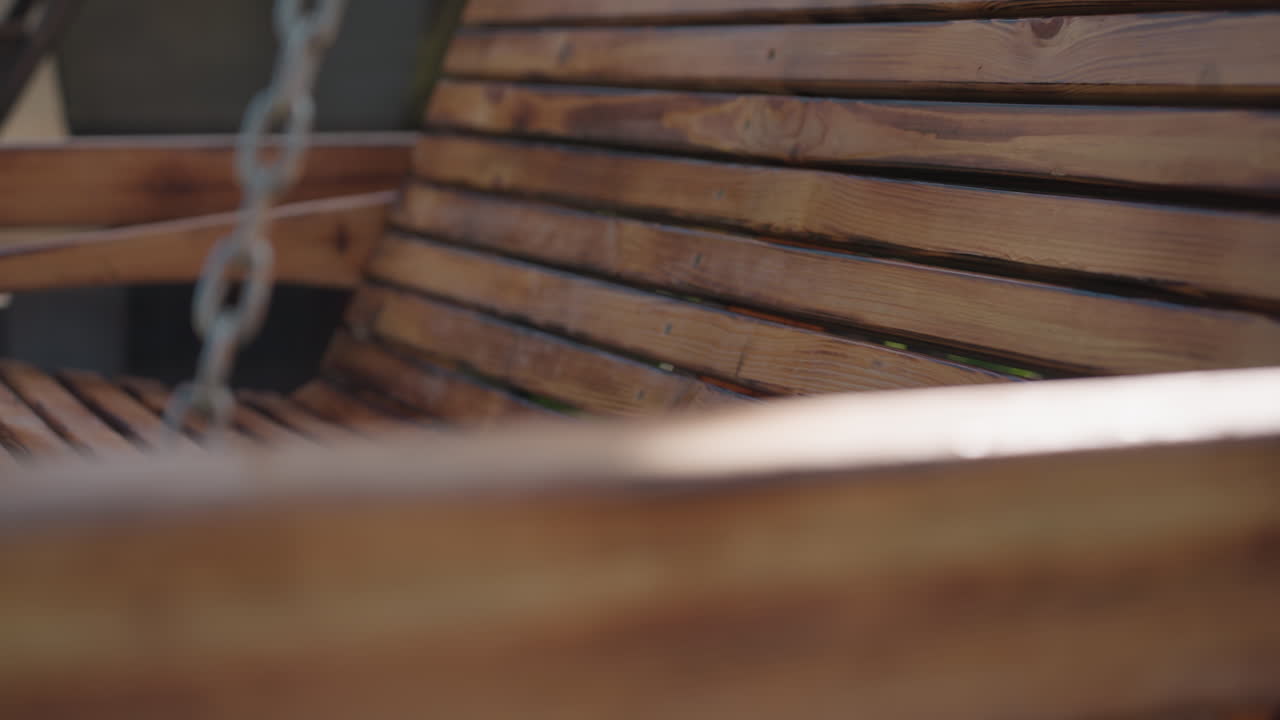 Close up of polished wooden bench swing with hanging metal chain in backyard, sun casting warm highlights on curved slats and grain texture