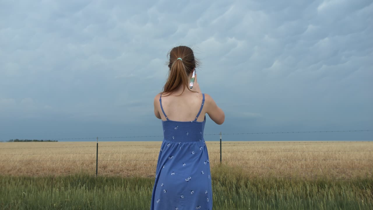 A young woman wearing a blue dress standing outside taking pictures on her phone of distant storm clouds.