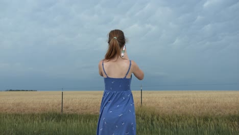 a young woman wearing a blue dress standing outside taking pictures on her phone of distant storm clouds.