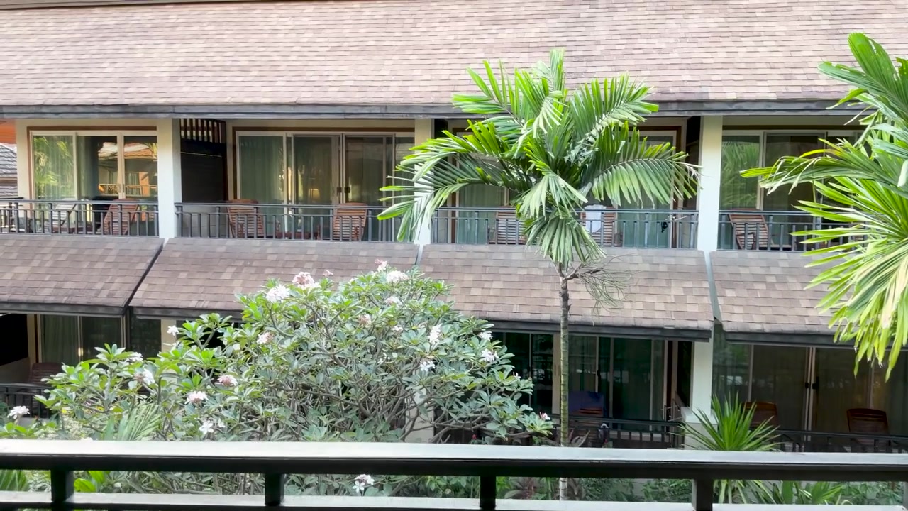 La telecamera si sposta da un letto con decorazioni tailandesi a un balcone che si affaccia sui giardini di un resort tropicale.