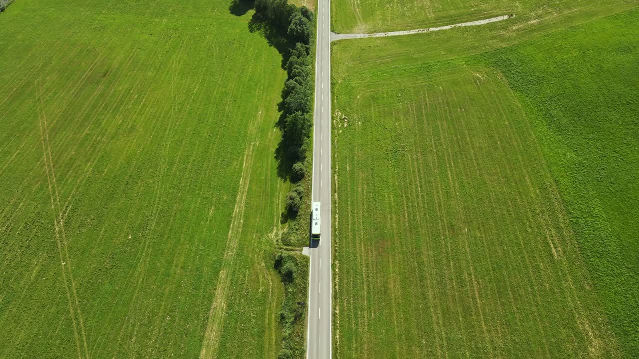 Aerial top-down view of a bus traveling along a straight rural road flanked by vast green fields. The scene highlights the simplicity and tranquility of countryside travel under bright daylight.