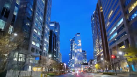 driving through a vibrant city at night, towering buildings glow against the dark sky while street lights reflect on the wet pavement, capturing the lively atmosphere of urban living.