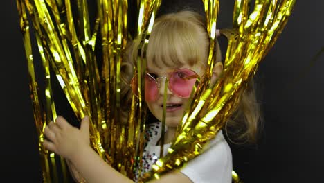 happy child jumping, playing, fooling around in shiny foil fringe golden curtain. little blonde girl