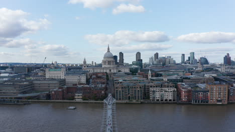 Descending-shot-of-Thames-river-waterfront-at-Millennium-bridge.-Saint-Pauls-Cathedral-with-large-dome-and-group-of-skyscrapers-in-background.-London,-UK