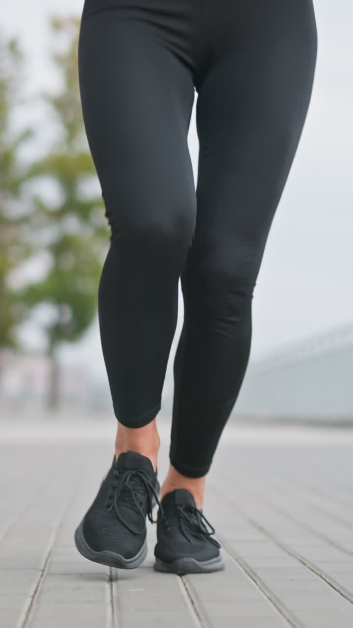 Close-up of woman s legs in black leggings and sneakers jogging in outdoor path with blurred background of trees, poles, and iron fence, showcasing fitness and active lifestyle