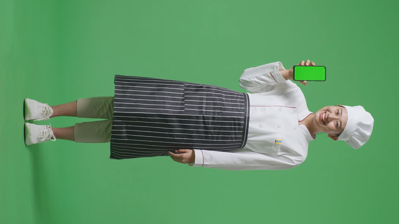 Full Body Of Asian Woman Chef Smiling And Showing Green Screen Smartphone To The Camera While Standing In The Green Screen Background Studio