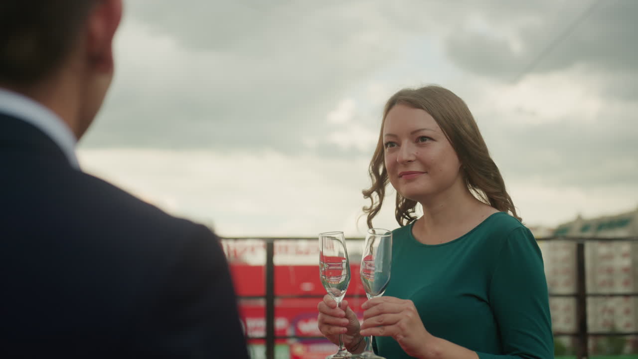 brindisi su un tetto con una donna bianca che tiene in mano dei flute di champagne, sorridendo verso il partner con lo skyline nuvoloso della città sullo sfondo, vestito verde, brezza leggera, conversazione intima e atmosfera calda serale
