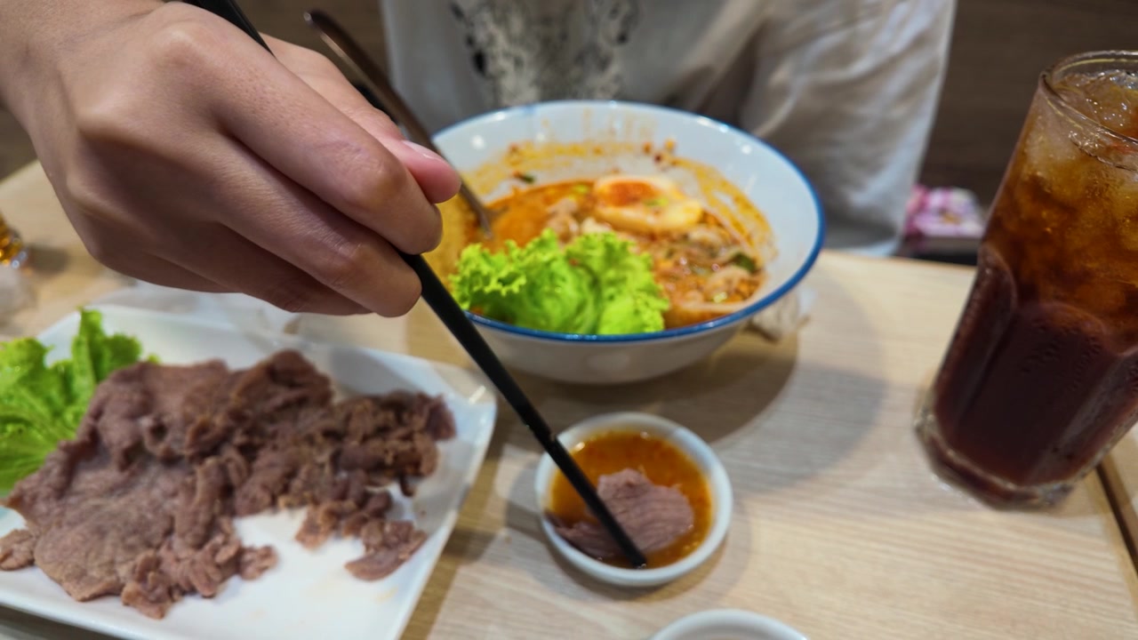 スパイシーな麺スープの横で、箸を使って牛肉をソースに浸している手のクローズアップ。