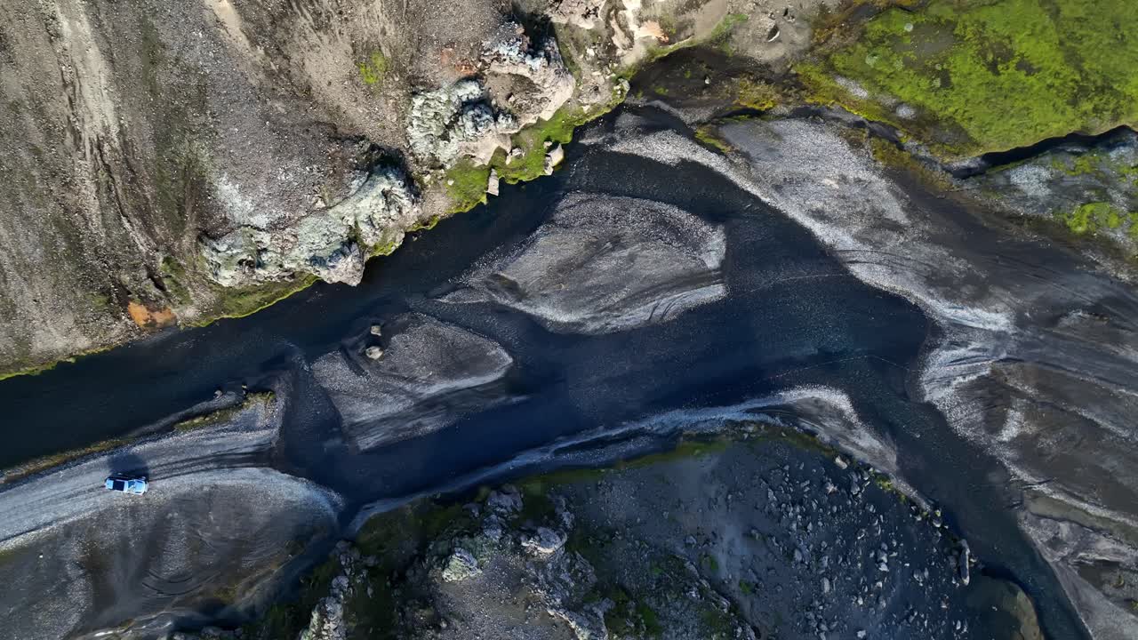 Aerial view of a 4×4 reversing along a gravel track beside a braided riverbed carved through dark volcanic sediment in the Icelandic highlands