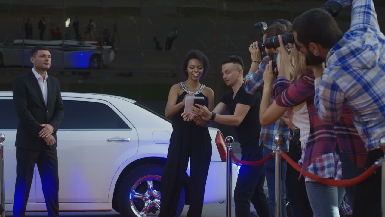 A celebrity signs autographs for paparazzi at a red carpet event with a limousine and security