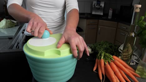 Woman-Using-Salad-Spinner-In-The-Kitchen-With-Pieces-Of-Fresh-Raw-Carrots-On-The-Side