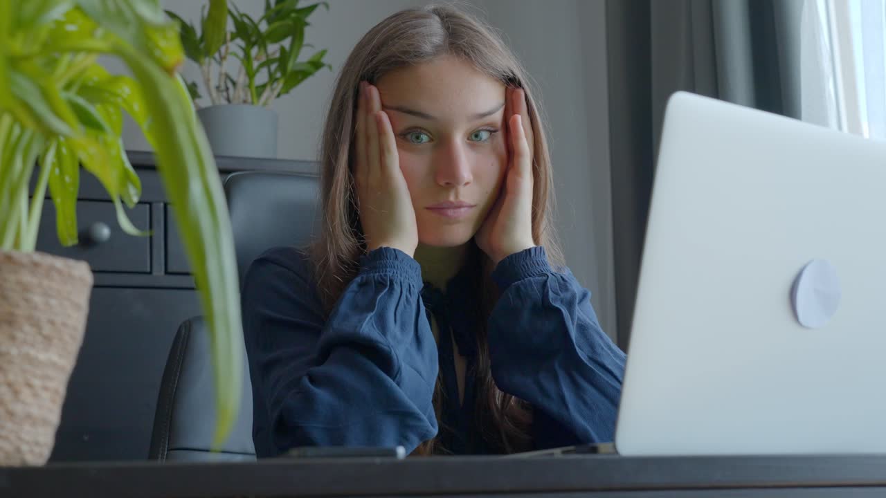 Stressed woman working on laptop looks at camera holding head with both hands