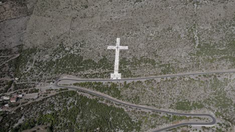 Video-De-Drones-De-Panorámica-Descriptiva-Sobre-La-Cruz-Blanca-Gigante-Al-Comienzo-Del-Paso-De-Montaña-De-La-Carretera-Sh20-En-Bridje,-Albania,-Se-Pueden-Ver-Las-Casas-Del-Pueblo,-Camino-Sinuoso