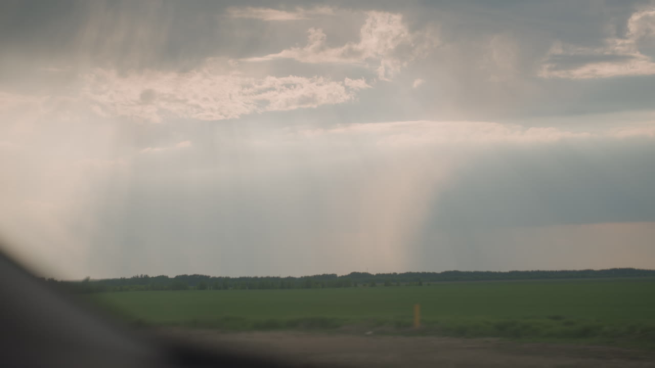 videographer captures serene rain shafts over green field under dramatic cloudy sky with sun beams piercing through clouds creating cinematic moody landscape