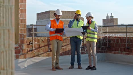 Mixed-races-male-and-female-builders-discussing-something-while-working-at-the-constructing-site-with-drafts,-documenrs-and-tablet-device.-Outdoors.