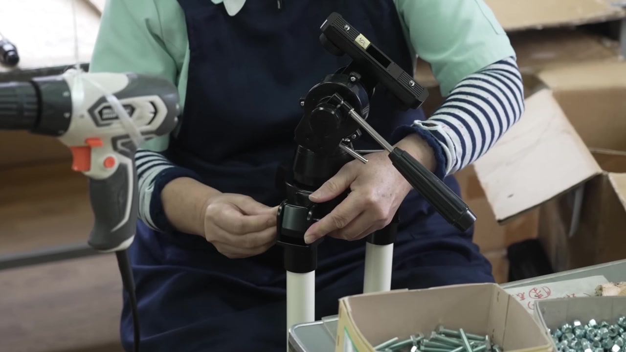 Worker assembling telescope tripod by attaching white legs to black equatorial mount head with hand tools