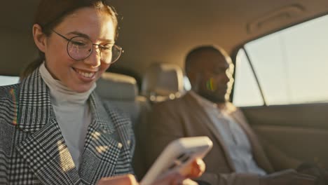 Close-up a brunette girl in round glasses and a gray jacket types on a white phone while traveling with her male colleague with Black skin in a modern car outside the city