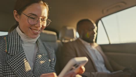 primo piano una ragazza bruna con gli occhiali rotondi e una giacca grigia scrive su un telefono bianco mentre viaggia con il suo collega di sesso maschile con la pelle nera in un'auto moderna fuori città