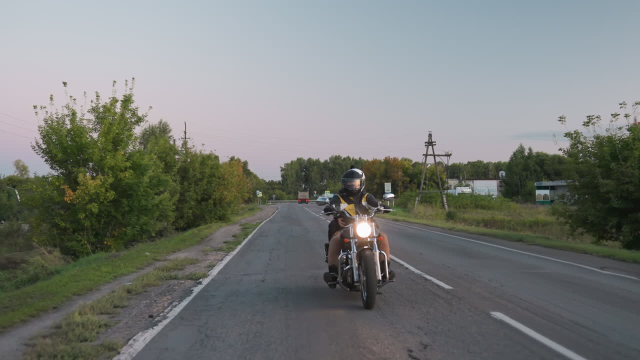 Motorbike rider wearing helmet drives on countryside road, trip, headlight shining while passing pedestrian signpost, distant car moves along opposite lane under clear evening sky