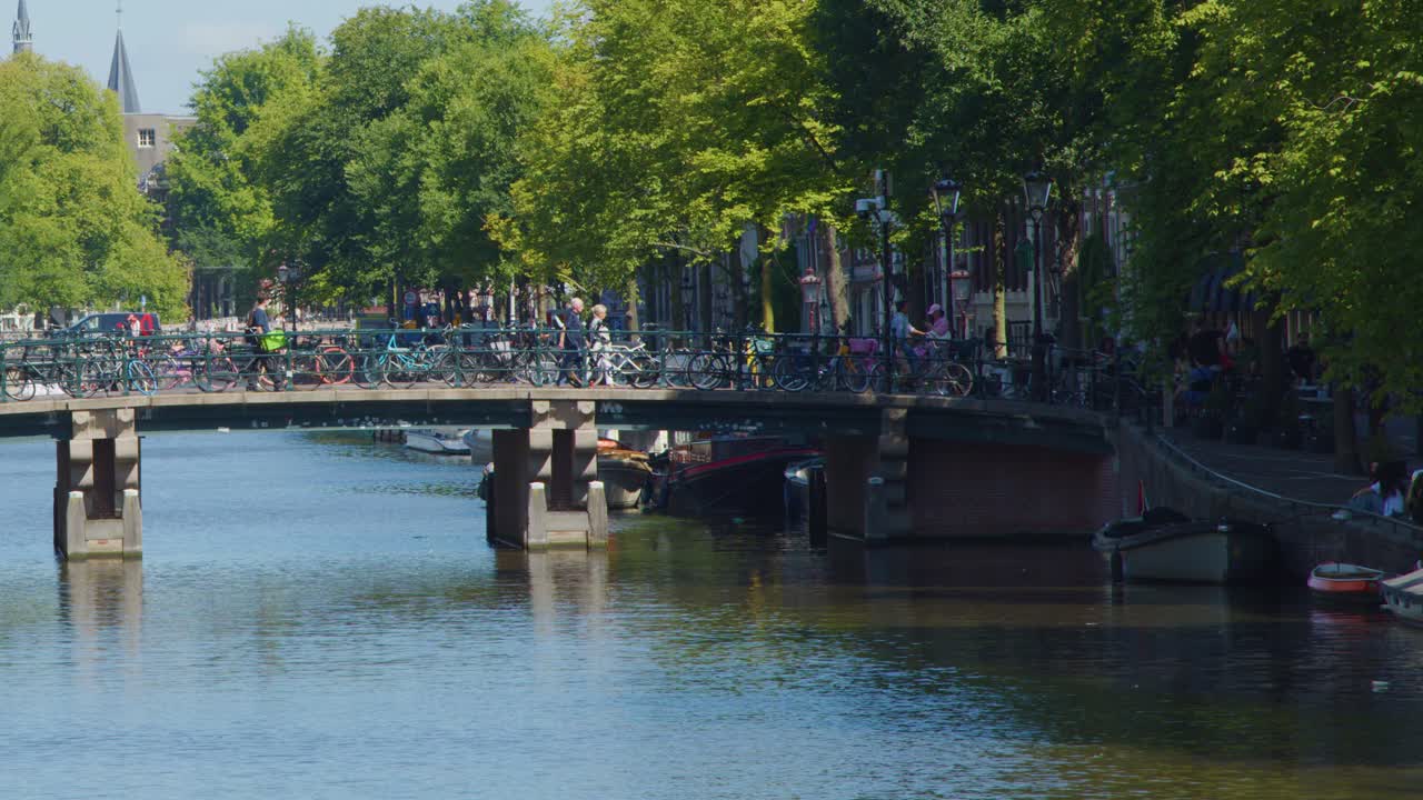 Cyclists cross a tree-lined bridge over a canal in Amsterdam, bright daylight, steady camera