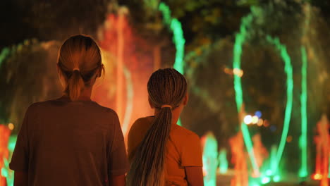 Madre-E-Hija-Admiran-La-Fuente-Con-Iluminación,-Un-Paseo-Por-La-Ciudad-Nocturna