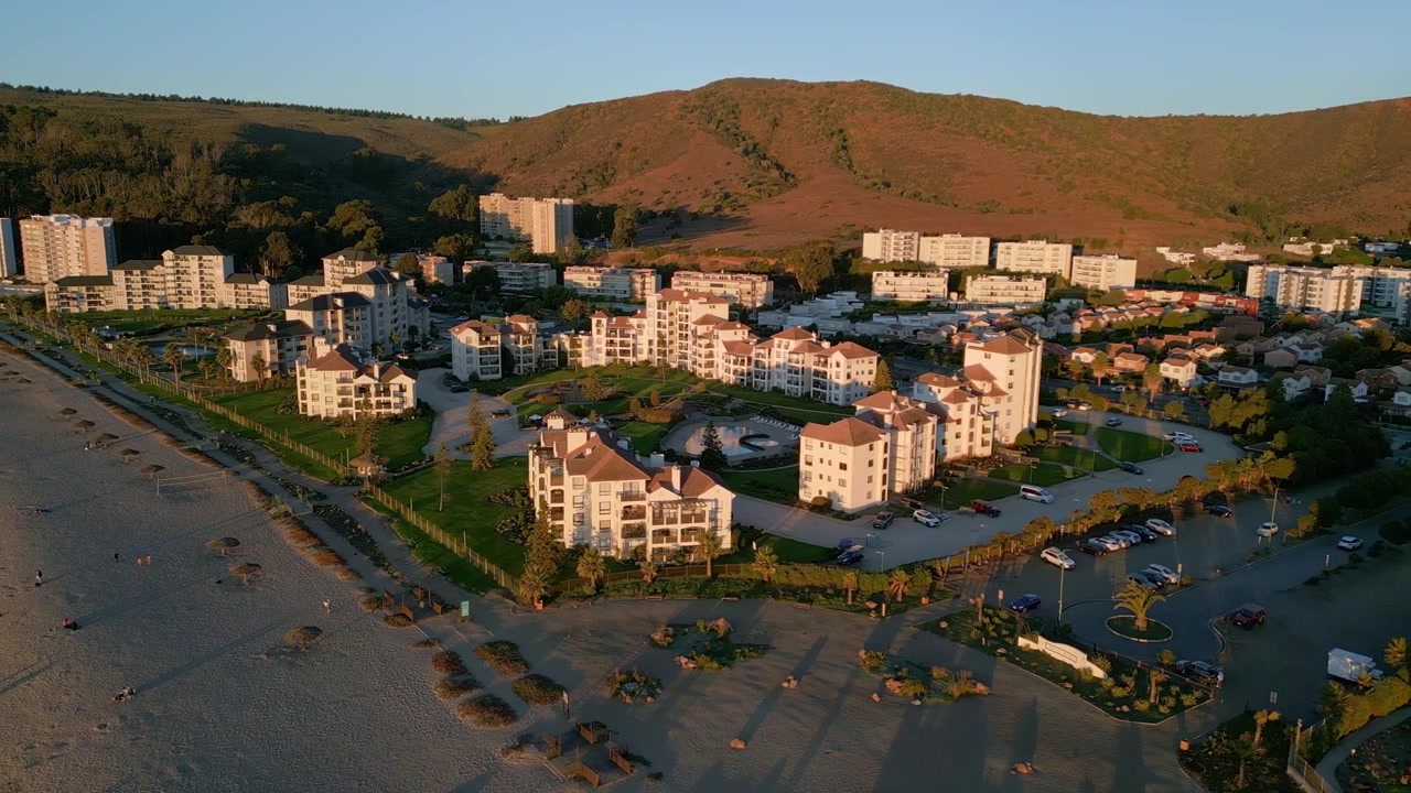 Panoramatický záběr na plážový resortní komplex se zelenými zahradami a bazénem zalitý teplým zlatým světlem západu slunce proti suchému zářícímu svahu. Punta Puyai, Papudo, Chile
